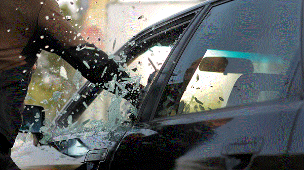 A man stands beside a smashed car, with shattered glass scattered around the vehicle.