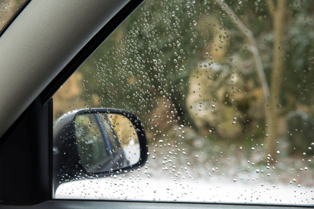 Raindrops on a car window, with windshield condensation, create a blurred view of the surroundings