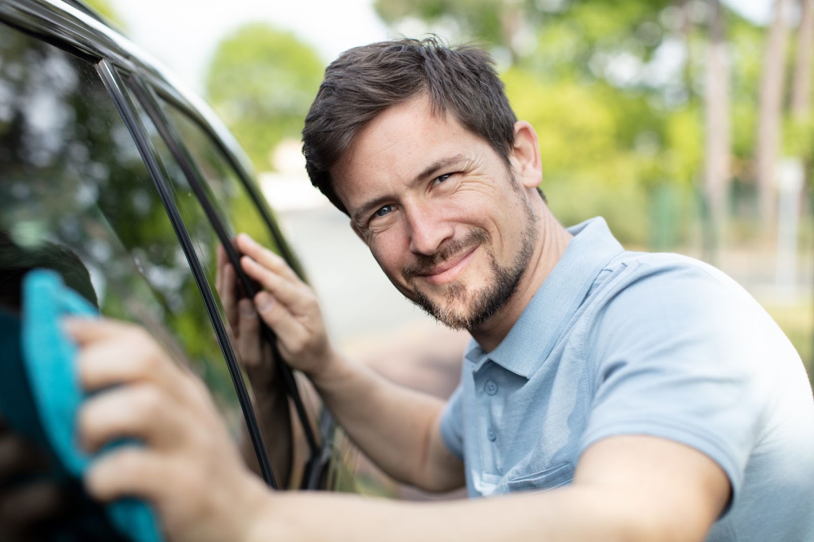 A man cleaning the window of his car, removing adhesive residue, with a windshield repair service in the background.