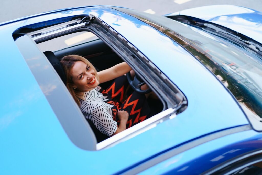 A woman seated in the driver's seat of a blue sports car, contemplating sunroof replacement options.