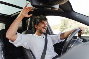 A man drives his car with a smile, waving cheerfully, showcasing a sunroof in need of replacement.